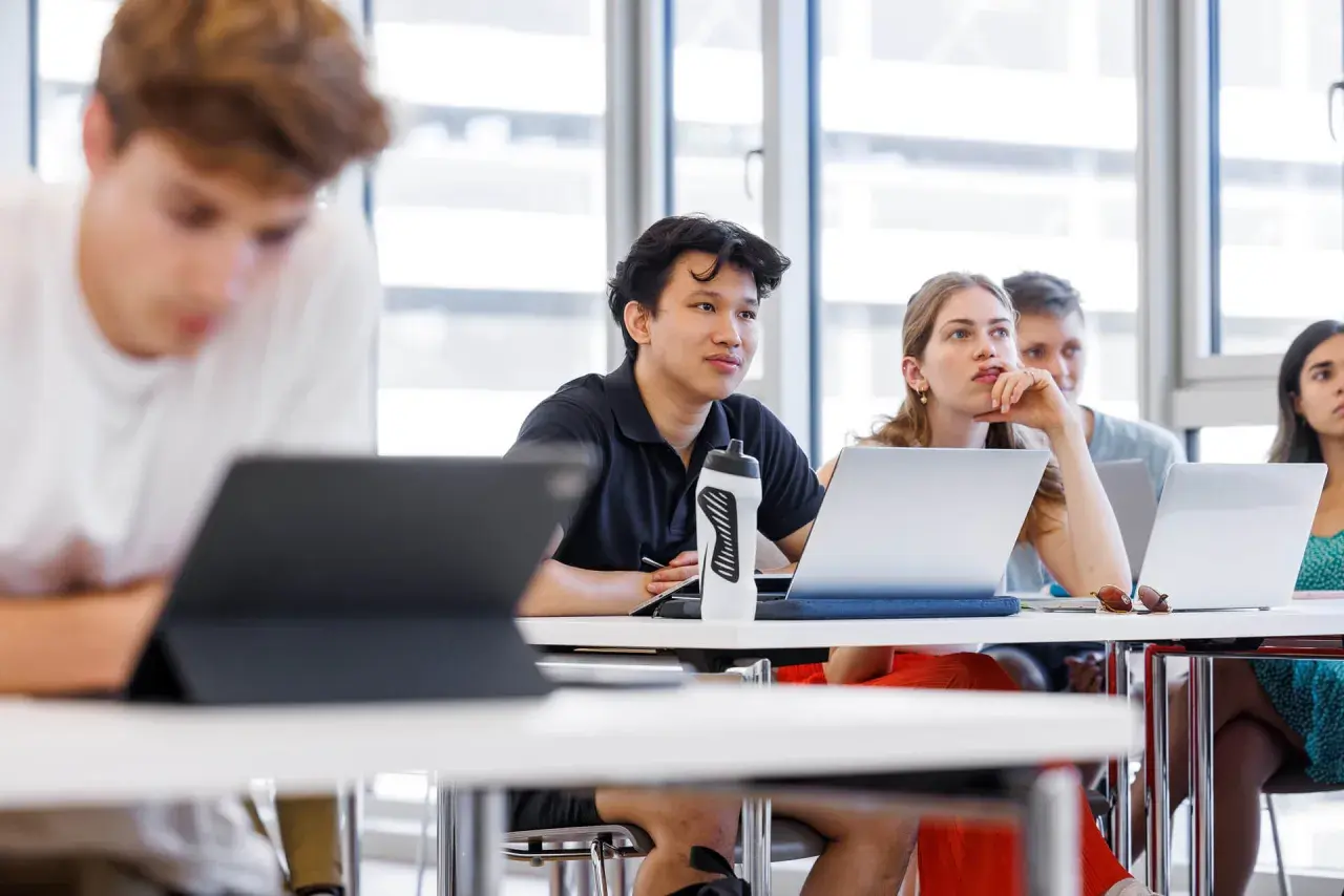 Studenten im Hörsaal, die sich auf ihre Lektionen konzentrieren. Ein junger Mann mit einem schwarzen Poloshirt und einer Wasserflasche, der aufmerksam zuhört. Dies ist ein typisches Bild für Management and Technology TUM.
