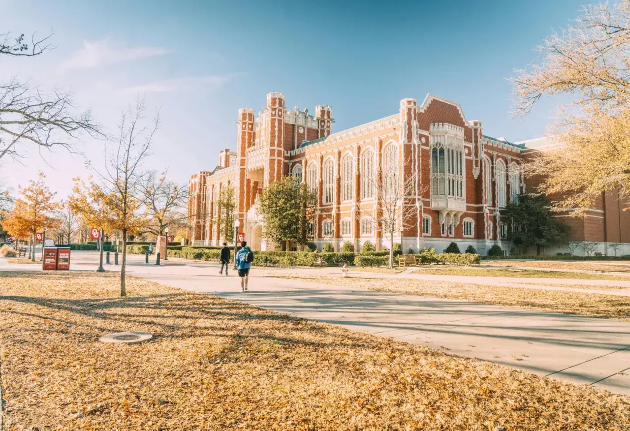 Herbstliche Szene auf einem Campus in den USA, mit Studenten, die an einem Backsteingebäude vorbeigehen. Ein schöner **college usa vergleich deutschland**-Moment.