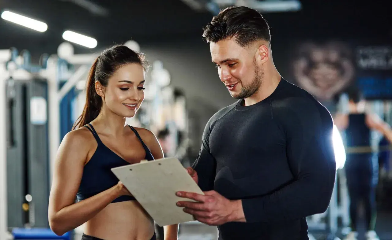 Ein Trainer bespricht mit einer Kundin Trainingspläne. Das Gehalt eines Sport- und Fitnesskaufmanns nach der Ausbildung ist attraktiv.