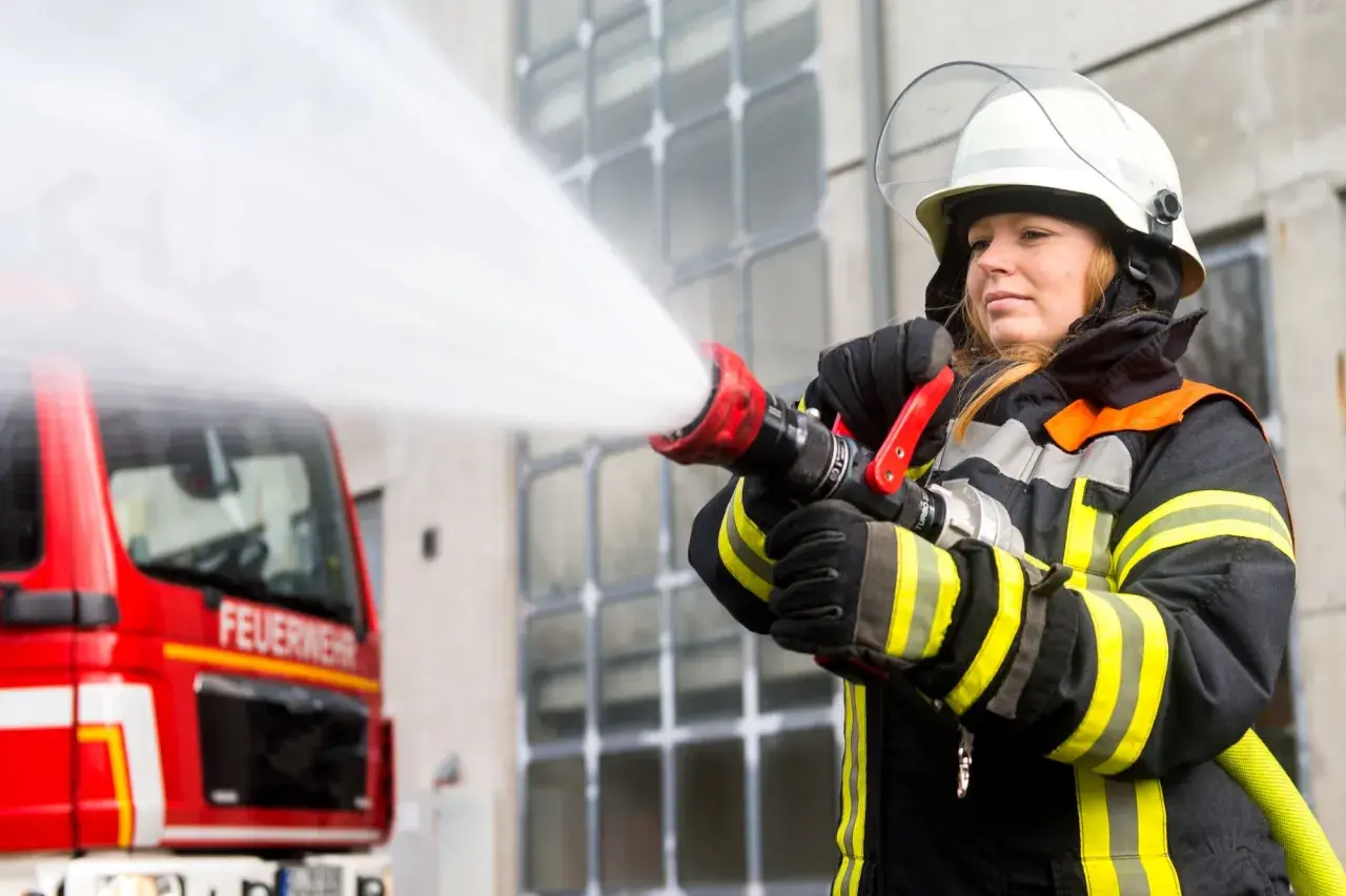 Eine Feuerwehrmann Frau löscht mit einem Schlauch einen Brand. Das rote Feuerwehrauto steht im Hintergrund.