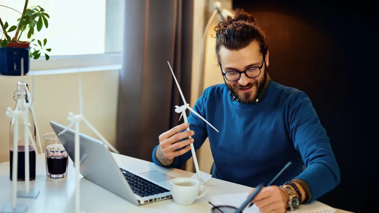 Ein Mann mit Brille und Bart hält ein Modell einer Windkraftanlage. Er studiert es, während er Notizen macht. Ein Laptop und eine Pflanze stehen auf dem Tisch. Ein angehender Wirtschaftsingenieur im Bereich erneuerbare Energien.