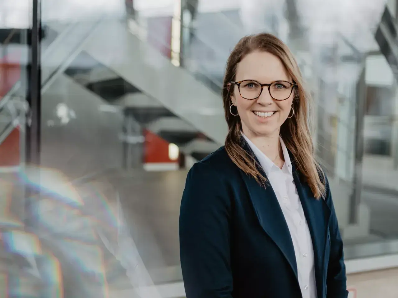 Lächelnde Frau mit Brille und Blazer vor moderner Glasfassade, inspiriert vom Studium.