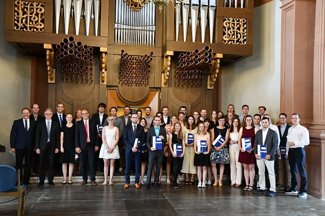 Absolventen mit Urkunden vor einer Orgel, die ihr erfolgreiches Diplom Studium feiern.