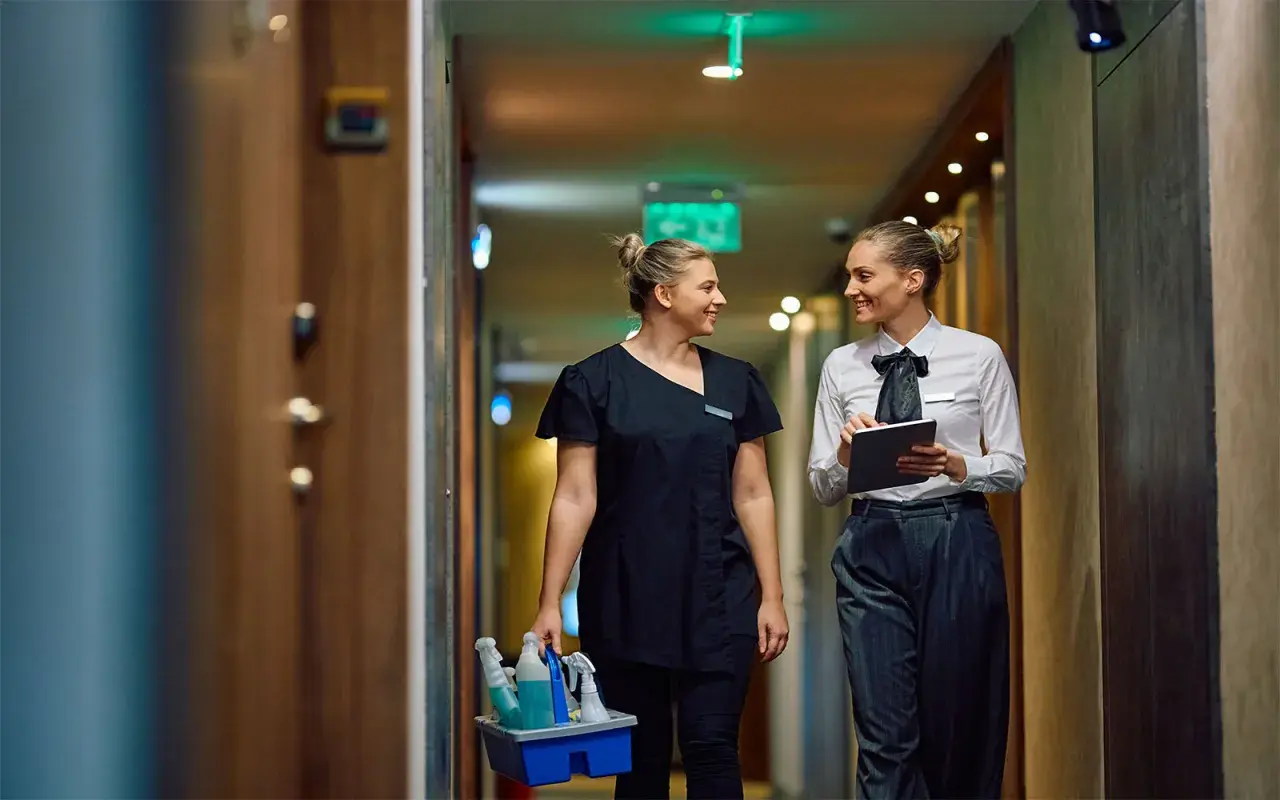 Zwei Frauen im Hotelgang, eine mit Reinigungsmitteln, die andere mit Tablet. Sie sprechen über das Hauswirtschafterin Gehalt.