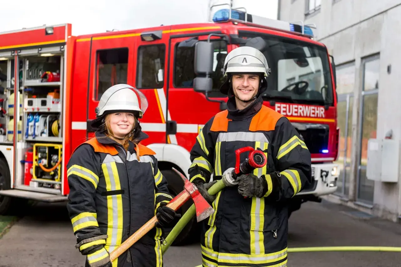 Zwei Feuerwehrleute mit Helm und Schutzkleidung stehen vor einem roten Feuerwehrauto. Sie halten Werkzeuge bereit. So kommt man zur Berufsfeuerwehr.