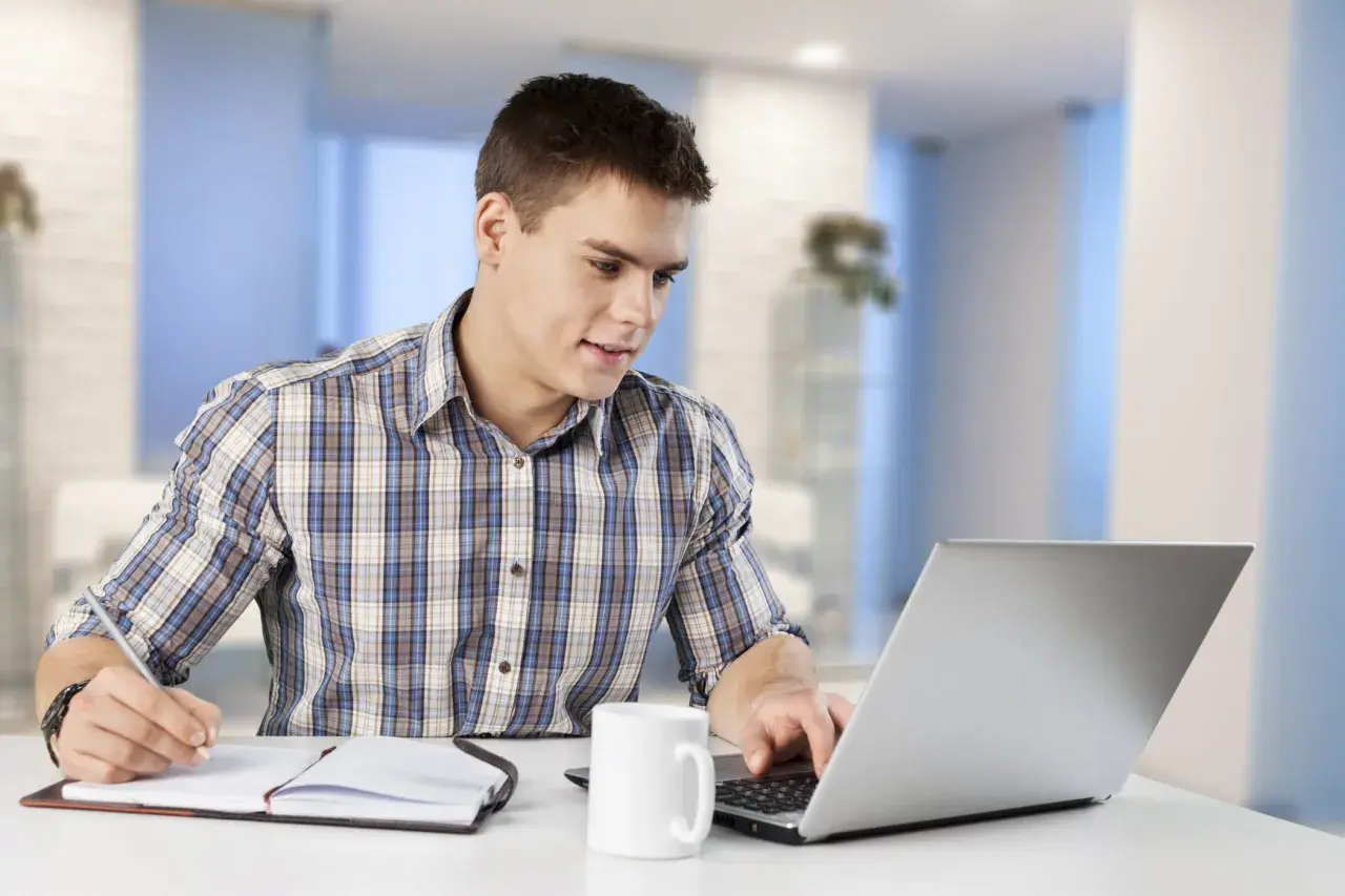 Junger Mann am Laptop, Notizen machend. Er informiert sich über das kaufmann e commerce gehalt.
