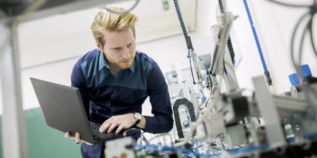 Ein junger ingenieur mit Bart arbeitet konzentriert an einem komplexen mechanischen Aufbau und bedient dabei einen Laptop.
