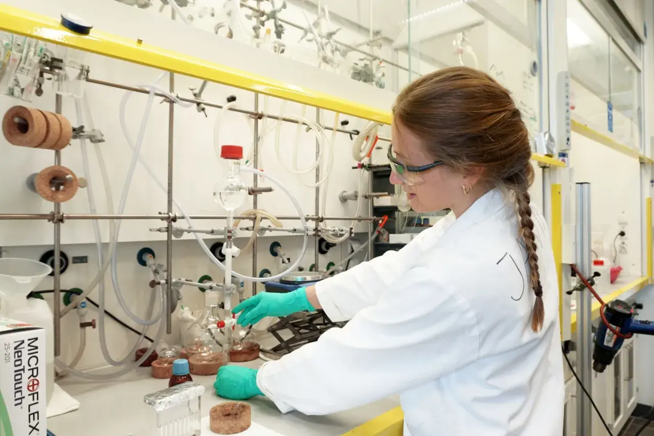 Junge Frau im Labor mit Schutzbrille und Handschuhen, die an einem Experiment arbeitet. Ein Schritt auf dem Weg zum Promotion Studium.