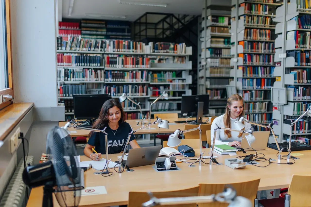 Zwei Studentinnen lernen in einer Bibliothek. Die Bedeutung von Bildung wird hier durch konzentriertes Arbeiten an Laptops und Büchern promoviert.