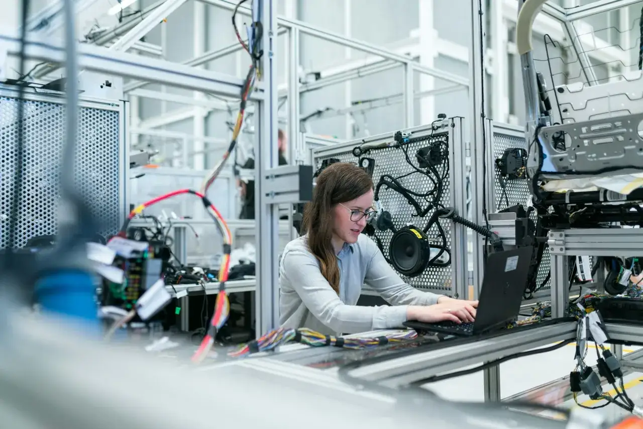 Ingenieurin arbeitet an einem Laptop in einem modernen Werk, umgeben von komplexen Kabelbäumen und technischen Komponenten.