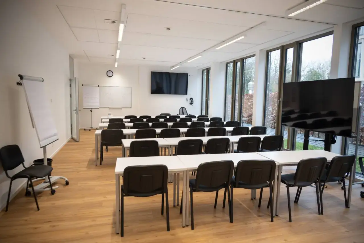 Leerer Seminarraum mit Reihen von Tischen und Stühlen, Flipchart und großen Bildschirmen. Perfekt für Schulungen am iu Bonn.