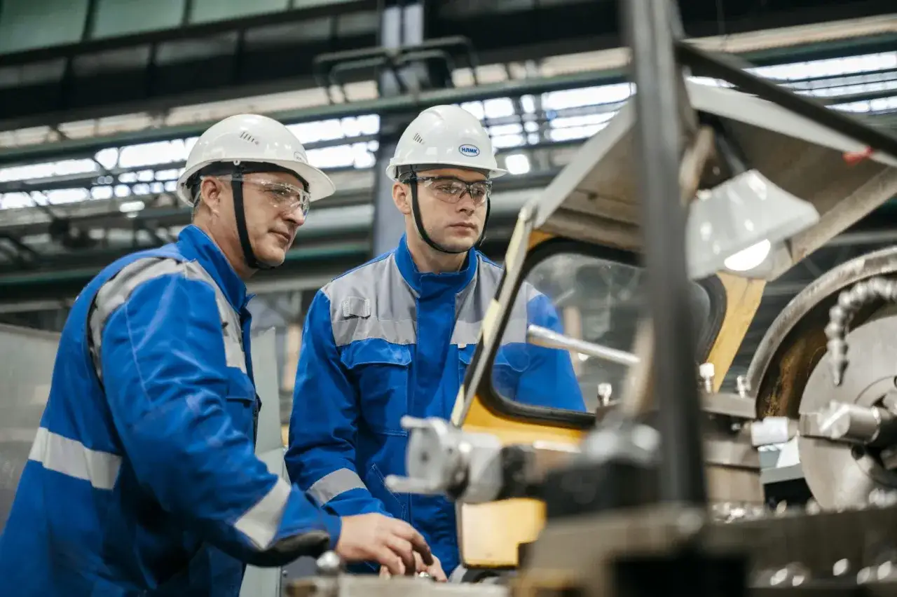 Zwei M&auml;nner in blauen Arbeitsanz&uuml;gen und Schutzhelmen arbeiten an einer Maschine. Sie sind Teil der industrie berufe, die das R&uuml;ckgrat der Produktion bilden.