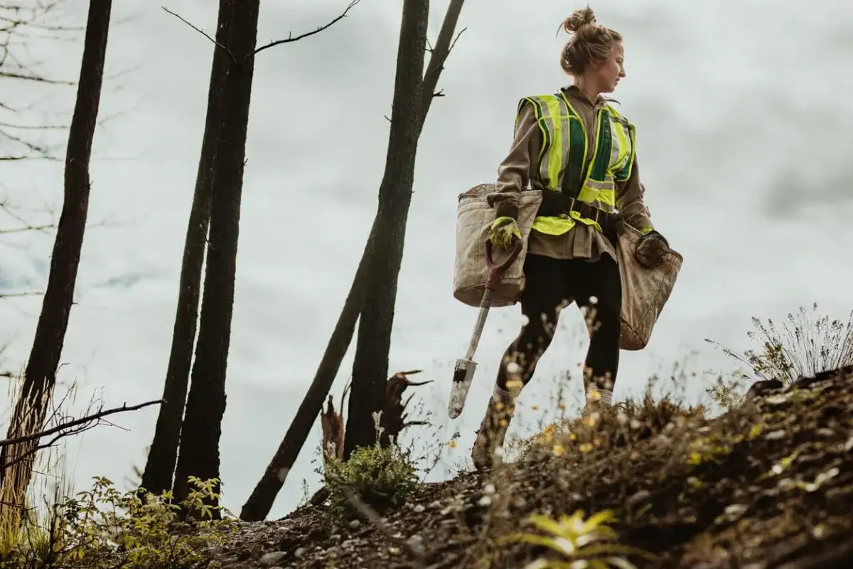Junge Frau in Warnweste mit Spaten und Pflanzbeutel im Wald. Die Ausbildung zum Forstwirt bietet spannende Arbeit und ein gutes Gehalt.