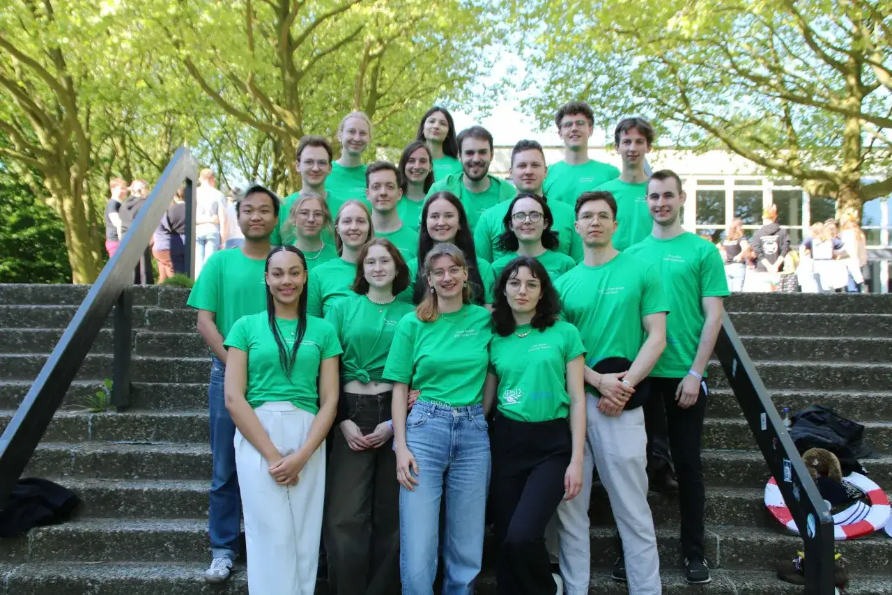 Eine Gruppe junger Leute in grünen T-Shirts, die für ihr Studium posieren, steht auf einer Treppe unter Bäumen.