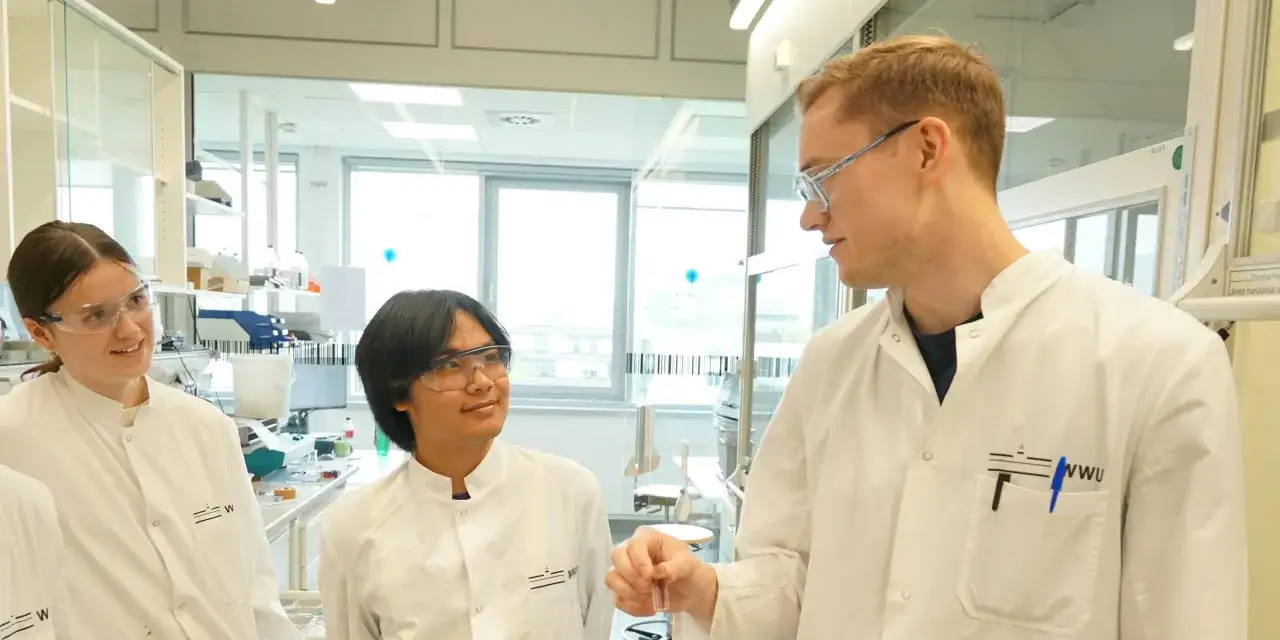Drei Studierende im weißen Kittel diskutieren im Labor. Ein Mann hält ein Reagenzglas. Ihr Pharmazie Studium NRW beginnt hier.