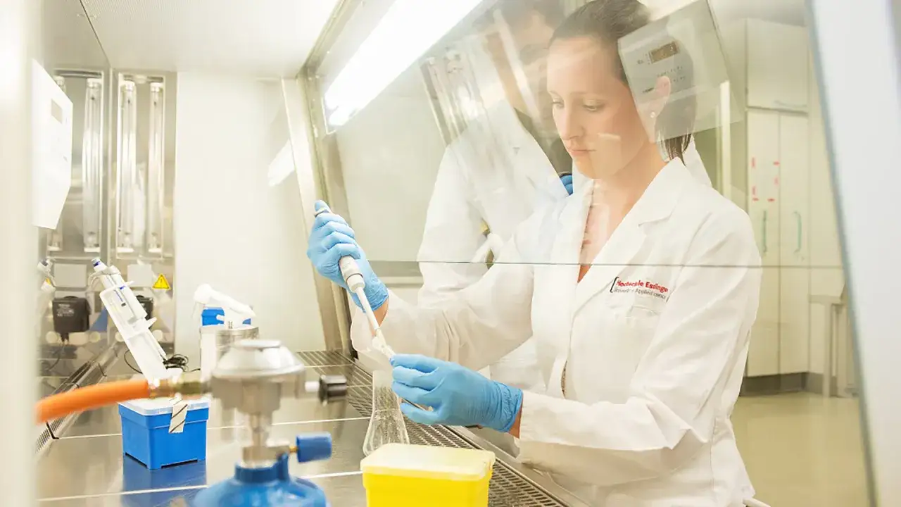 Frau im Labor, die mit einer Pipette arbeitet. Ein Kollege im Hintergrund. Studium Biotechnologie.