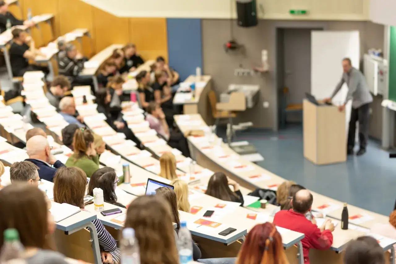 Vorlesungssaal mit vielen Studierenden, die aufmerksam einem Vortragenden zuhören. Ein typischer Ort, um mehr über das zu lernen, was ein Bachelor ist.