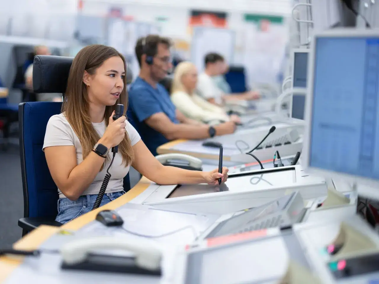 Eine Frau spricht ins Mikrofon und bedient ein Touchpad. Sie ist Teil eines Teams, das im Hintergrund arbeitet. Das Bild zeigt, was ein Fluglotse tut: die Sicherheit im Luftverkehr gew&auml;hrleisten.
