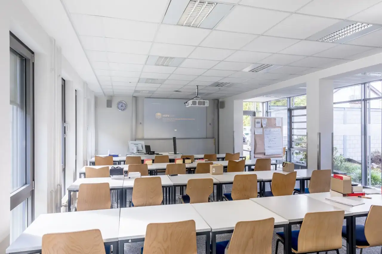 Leerer Hörsaal der HSPV NRW mit Reihen von Tischen und Stühlen. Eine Leinwand mit dem Logo der Hochschule ist an der Wand.