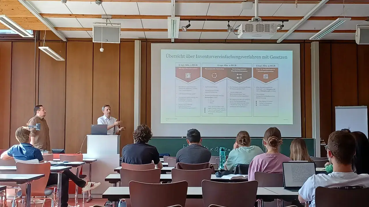Dozent erklärt im Hörsaal die Übersicht über Inventurvereinfachungsverfahren mit Gesetzen, relevant für das Steuerrecht. Studierende hören zu.