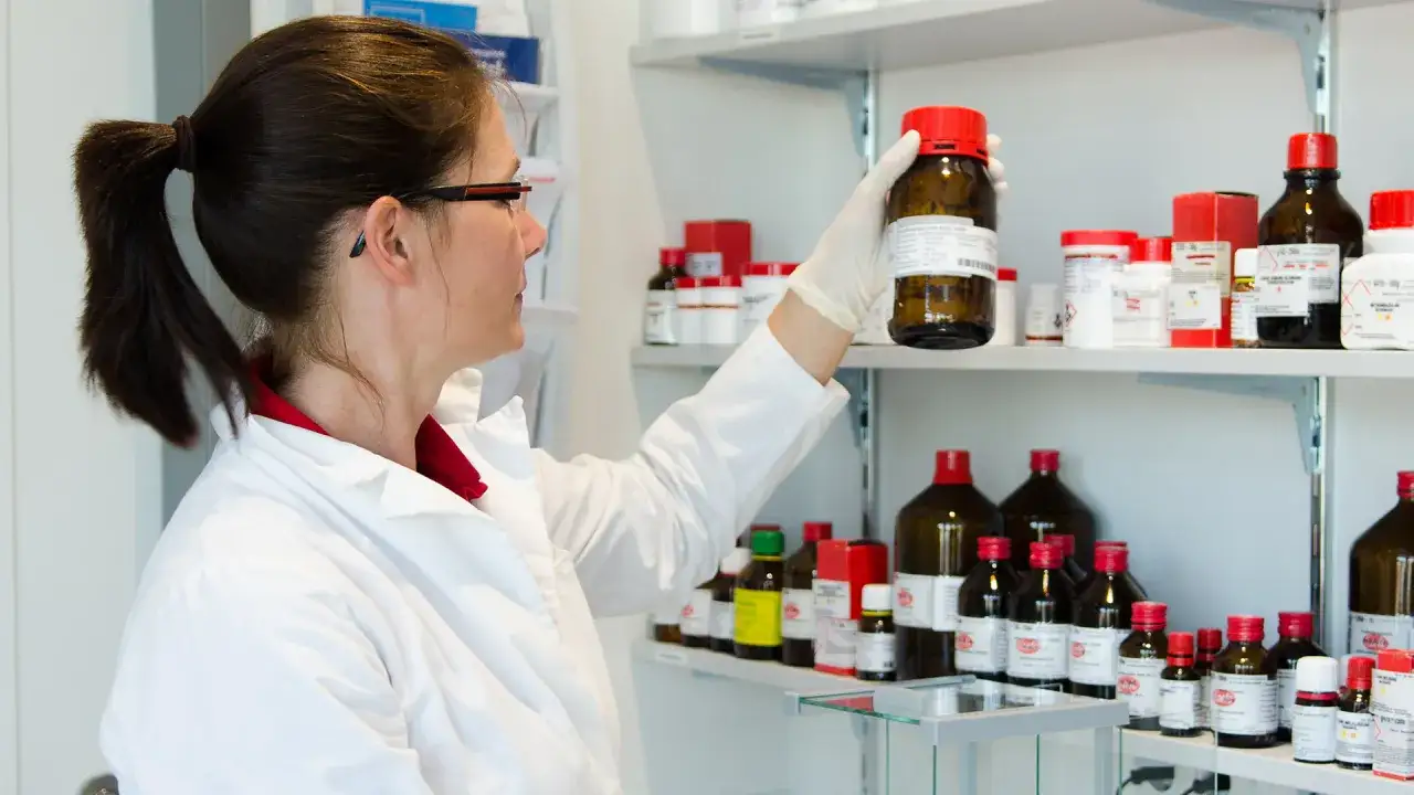 A woman in a lab coat examines a bottle on a shelf filled with chemicals. She's preparing for her state exam, wondering,
