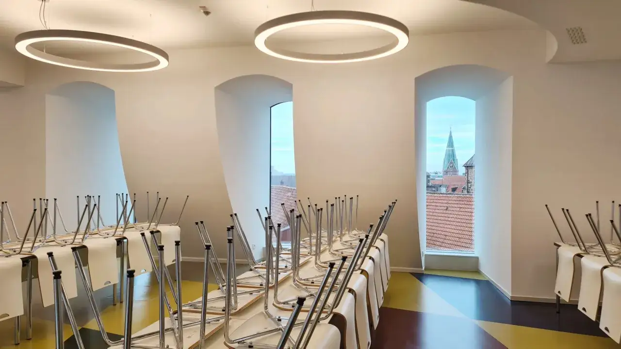Leere Tische und gestapelte Stühle in einem hellen Raum mit großen Fenstern, die einen Blick auf eine Stadt mit Kirchturm freigeben. So könnte ein Teil eines modernen Campus aussehen.