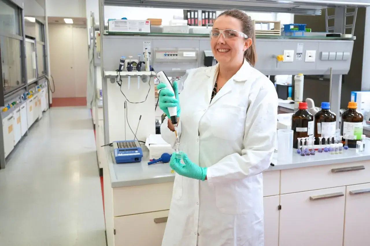 Frau in Laborkittel und Schutzbrille arbeitet an ihrer Masterarbeit mit Pipette und Reagenzgläsern in einem modernen Labor.